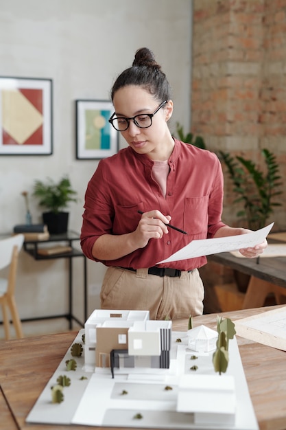 Selbstbewusste junge hispanische Frau in Brille, die am Schreibtisch mit Maquette des modernen Gebäudes steht und den Hausplan analysiert