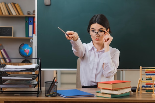 Selbstbewusste junge Lehrerin, die eine Brille trägt, zeigt an der Seite mit einem Zeiger, der mit Schulwerkzeugen im Klassenzimmer am Schreibtisch sitzt