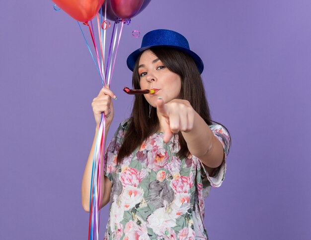 Selbstbewusste junge schöne frau mit partyhut, die luftballons hält und die partypfeife bläst, die ihnen geste isoliert auf blauer wand zeigt