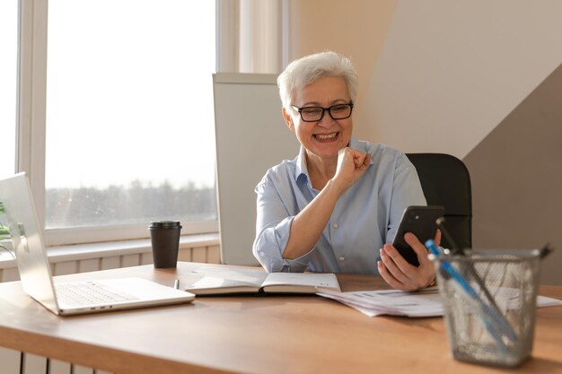 Selbstbewusste stilvolle europäische ältere Frau mittleren Alters, die am Arbeitsplatz ein Smartphone benutzt, stilvoll älter