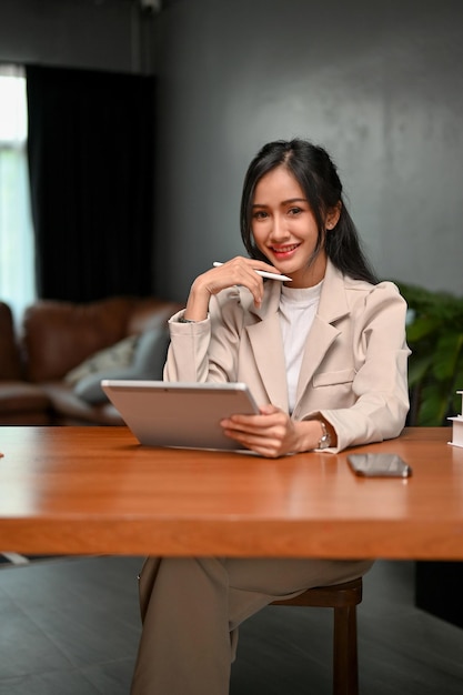 Selbstbewusste tausendjährige asiatische Geschäftsfrau sitzt mit ihrem digitalen Tablet an ihrem Schreibtisch