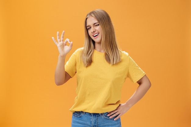 Selbstbewusste und geschickte Studentin, die der Lehrerin versichert, dass sie großartige Arbeit leistet, die Hand an der Taille hält, mit selbstbewusstem Blick in die Kamera zwinkert und eine gute Geste zeigt, die sicherstellt, dass über der orangefarbenen Wand alles in Ordnung ist