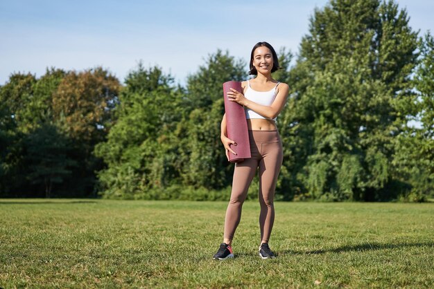 Selbstbewusste und sportliche junge Asiatin, die im Park steht und eine lächelnde Sportlerin mit Gummimatte trägt