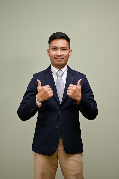 Selbstbewusster asiatischer Geschäftsmann, der vor einem grünen Studiohintergrund steht und Daumen hoch zeigt