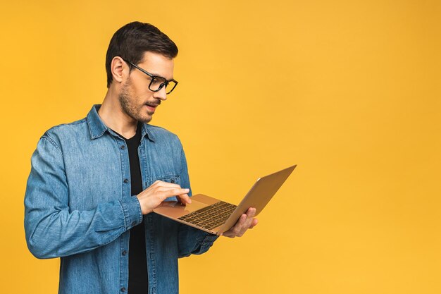 Selbstbewusster Geschäftsexperte Selbstbewusster junger, gutaussehender bärtiger Mann in lässigem Laptop und lächelnd, während er vor isoliertem gelbem Hintergrund steht