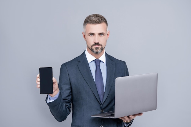 Selbstbewusster Geschäftsmann im geschäftsmäßigen Anzug hält Computer und präsentiert Smartphone, Werbung.