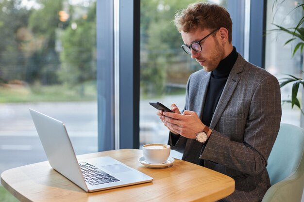 Selbstbewusster Geschäftsmann mit Handy sitzt am Schreibtisch mit Laptop bei einer Tasse Aromakaffee im Café