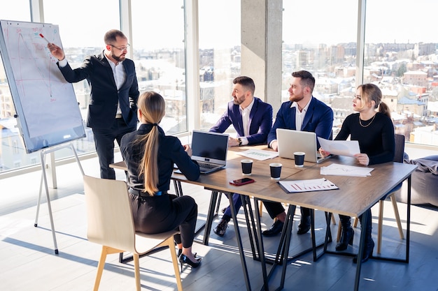 Selbstbewusster Geschäftsmann präsentiert bei einer Firmenversammlung ein neues Projekt im Sitzungssaal. Schöne Auditoren unterhalten sich mit verschiedenen Partnern über das Geschäft, indem sie ein Whiteboard und Grafiken verwenden.