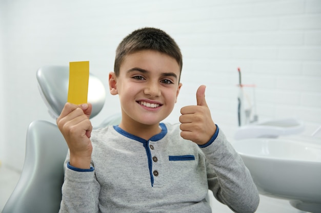 Selbstbewusster, gutaussehender Kinderjunge hält eine gelbe Visitenkarte mit Kopien-Werberaum, zeigt den Daumen nach oben und lächelt mit einem schönen zahnigen Lächeln, das die Kamera anschaut, während er nach der Zahnbehandlung im Zahnarztstuhl sitzt