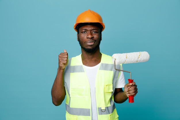 Selbstbewusster junger afrikanisch-amerikanischer Baumeister in Uniform mit Walzenbürste isoliert auf blauem Hintergrund