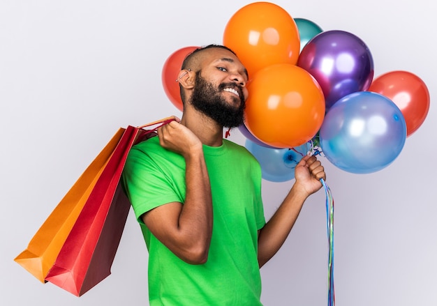 Selbstbewusster junger Afroamerikaner mit Partyhut, der Luftballons hält und Geschenktüte auf die Schulter legt