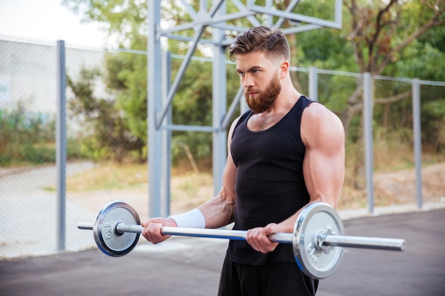 Selbstbewusster junger bärtiger Fitness-Mann, der draußen Übungen mit der Langhantel macht