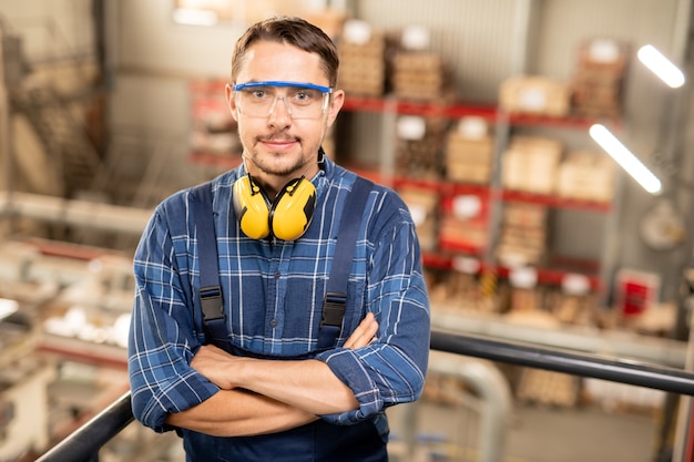 Selbstbewusster junger, bewaffneter Ingenieur einer großen modernen Fabrik, der Sie durch eine Schutzbrille ansieht