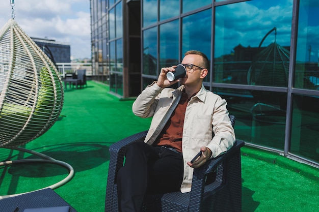 Selbstbewusster junger Geschäftsmann in formeller Kleidung und stilvoller Brille ruft ein Smartphone an, während er mit Kaffee auf der Terrasse eines modernen Bürogebäudes mit Glaswand sitzt