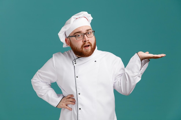 Selbstbewusster junger männlicher Koch mit Brille, Uniform und Mütze, der in die Kamera blickt und eine leere Hand zeigt, die eine andere Hand auf der Taille isoliert auf blauem Hintergrund hält