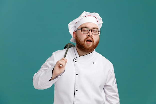 Selbstbewusster junger männlicher Koch mit einheitlicher Brille und Kappe, die eine Schöpfkelle auf der Schulter hält und isoliert auf blauem Hintergrund in die Kamera blickt