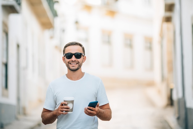 Selbstbewusster junger Mann mit Brille, der eine Kaffeetasse hält und sein Smartphone benutzt, während er im Freien spazieren geht