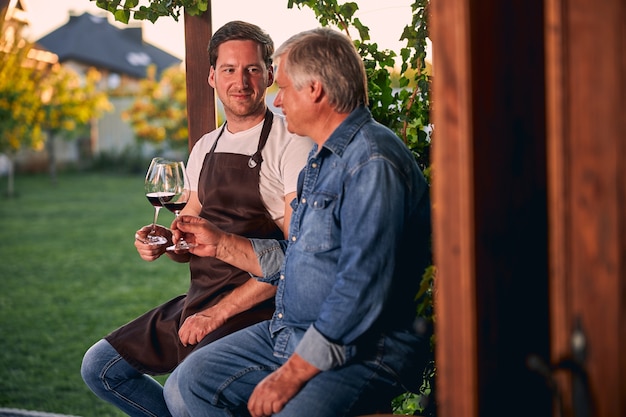 Selbstbewusster junger Sommelier, der einem reifen Mann lächelt, während er mit ihm im Freien sitzt. Gläser Rotwein in den Händen