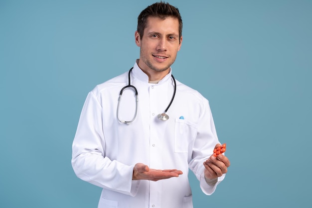 Selbstbewusster männlicher Arzt in weißer medizinischer Jacke, der Pillen hält, während er isoliert auf blauem Hintergrund in die Kamera blickt. Medizin-Konzept