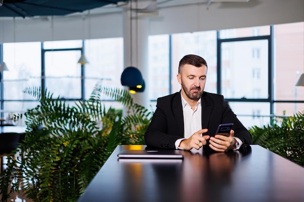 Selbstbewusster männlicher Geschäftsmann in einem Business-Anzug sitzt an einem Tisch mit einem Telefon und arbeitet aus der Ferne