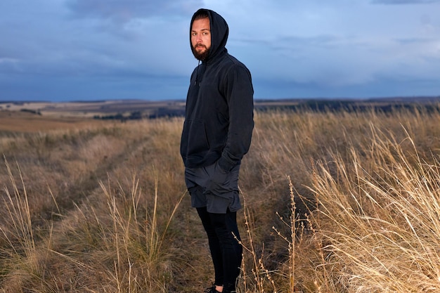 Selbstbewusster Mann, der sich die Kamera ansieht und sich nach dem Joggen ausruht, junger Mann in schwarzer Sportbekleidung, der allein auf dem Feld steht und die Hände in der Tasche hält. in der Natur im Freien. Outdoor-Porträt