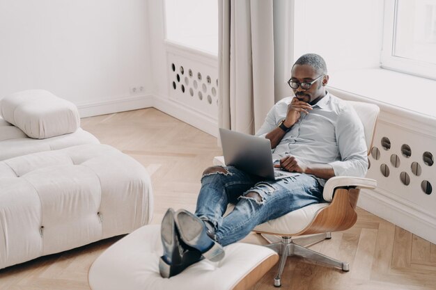 Selbstbewusster Profi bei der Arbeit vor dem Laptop Entspannter afrikanischer Geschäftsmann im gemütlichen Stuhl