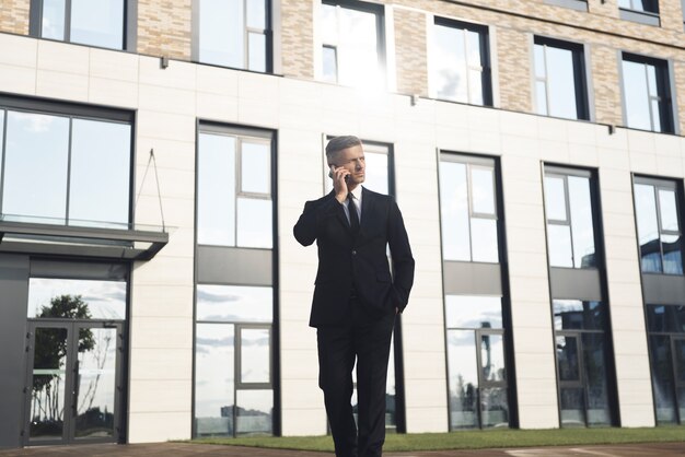 Foto selbstbewusster reifer geschäftsmann, der beim gehen vor dem bürogebäude telefoniert