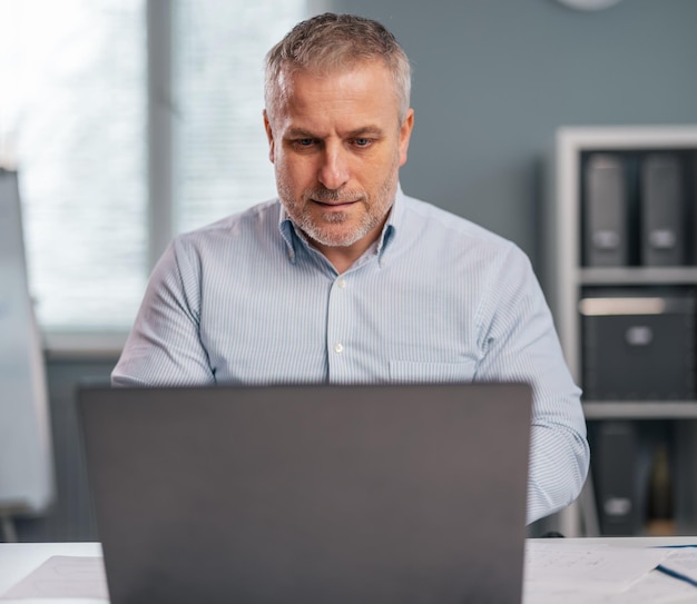 Selbstbewusster reifer Mann im formellen Hemd, der auf einem drahtlosen Laptop tippt, während er am Schreibtisch sitzt Kaukasische Geschäftsperson, die modernes Gadget für die Arbeit im Büro verwendet