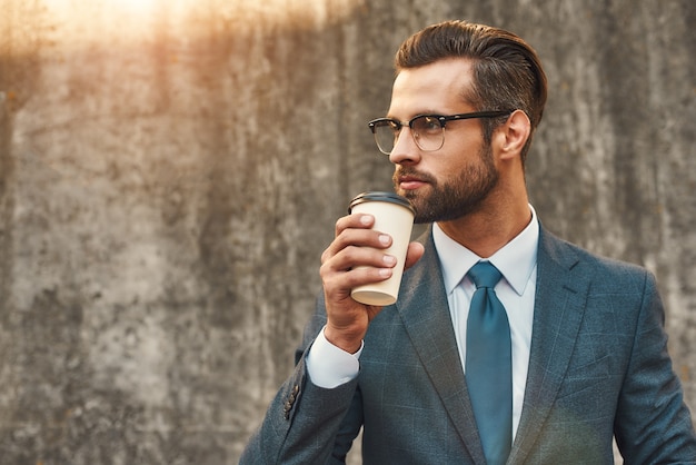 Selbstbewusster und erfolgreicher junger Geschäftsmann in formeller Kleidung und Brille, der Kaffee trinkt und schaut