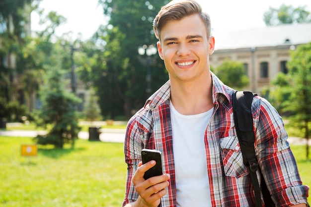 Selbstbewusster und erfolgreicher Schüler. Hübscher männlicher Student, der Handy hält und lächelt, während er im Freien steht