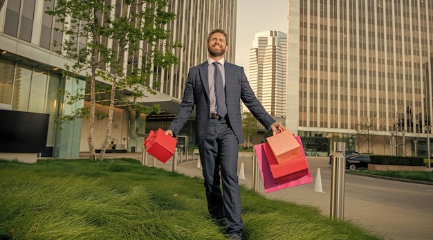 Selbstbewusster unternehmer im anzug mit einkaufstasche und geschenkbox außerhalb des bürorabatts