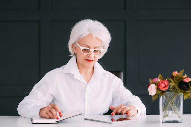 Selbstbewusstes Geschäftsporträt der gealterten Frau. Moderner Senioren-Lifestyle. Intelligente ältere Dame, die damit beschäftigt ist, Notizen im Tagesplaner zu machen, während sie das Tablet durchsucht.