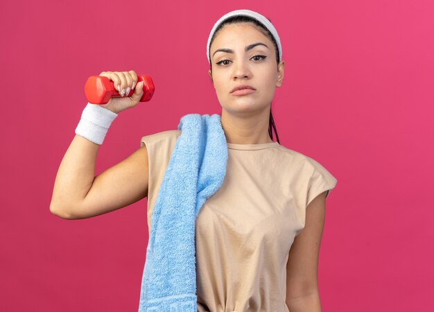 Selbstbewusstes junges kaukasisches sportliches Mädchen mit Stirnband und Armbändern, das Hantel mit Handtuch auf der Schulter anhebt und auf die Vorderseite isoliert auf rosa Wand schaut