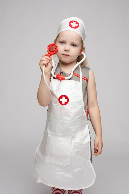 Selbstbewusstes kleines Mädchen, das im Uniformenmantel des Arztes posiert, der Stethoskop genießt, das Spielen genießt