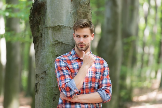 Selbstbewusstsein Kerl mit Borsten tragen kariertes Hemd lässig Mode Stil muskulöser Mann im Wald sexy Macho in aufgeknöpftem Hemd im Freien junger unrasierter Mann hat gepflegtes Haar männliche Schönheit und Mode