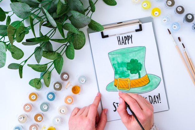 Selbstgemachte gemalte Grußkarte St. Patricks Day. Geschenkidee, Dekoration. DIY. Mach es selbst. Klee, Pinsel und Farbe. Weibliche Hände. St. Patricks Day-Hut.