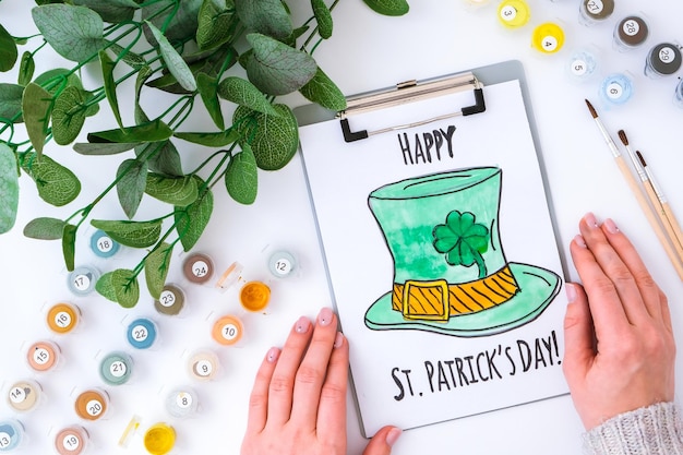 Selbstgemachte gemalte Grußkarte St. Patricks Day. Geschenkidee, Dekoration. DIY. Mach es selbst. Klee, Pinsel und Farbe. Weibliche Hände. St. Patricks Day-Hut.