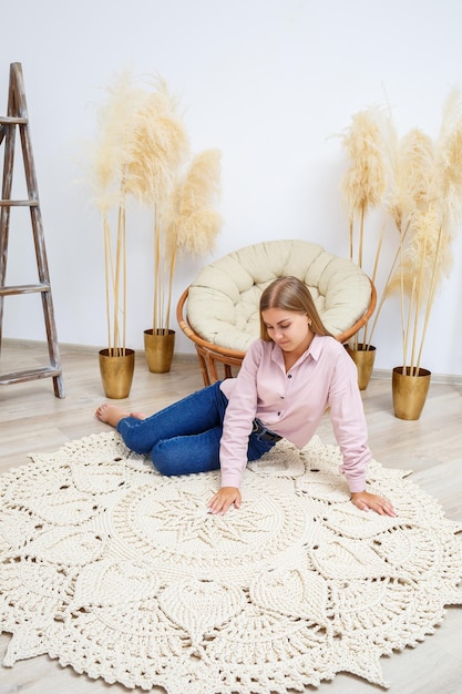 Selbstgemachter gestrickter Teppich aus dicken Fäden von Hand gefertigt Auf dem Boden liegt ein handgefertigter Teppich aus rosafarbenen Fäden, auf dem eine Frau mit einem Laptop in einem rosafarbenen Leinenhemd und Jeans sitzt