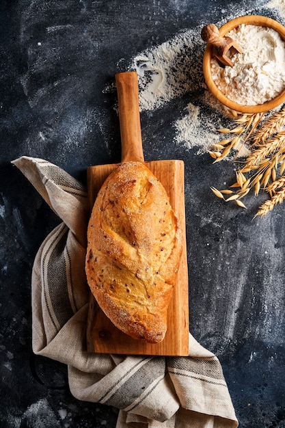 Selbstgemachtes Brot mit Samen auf einem Holzständer, Weizenmehl und Ohren auf grauem altem Betonhintergrundtisch.