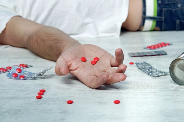 Selbstmord-, Depressions- und Überdosierungskonzept toter Mann mit einer Flasche Alkohol und Pillen, die nachts auf dem Tisch liegen, rote Tabletten in der Hand eines jungen Mannes