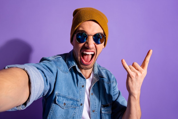 Selbstporträt des trendigen stilvollen fröhlichen unhöflichen schreienden Kerls, der selfie nimmt, das Sie gehörnte Finger-Rockzeichen trägt, das braunen Kappen-Denim trägt, der über lila lebendigem Farbhintergrund lokalisiert wird