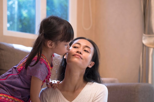 Selektiver Fokus auf das kleine asiatische Mädchen, das zu Hause im Wohnzimmer die Wange ihrer Mutter küsst