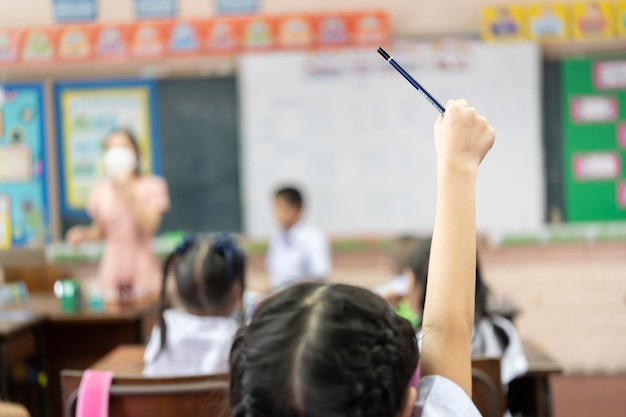 Selektiver Fokus auf die Hand Kinder oder Schulkinder oder Schüler, die Hände mit asiatischem Lehrer heben, der im Klassenzimmer in der Schule eine schützende Gesichtsmaske trägt