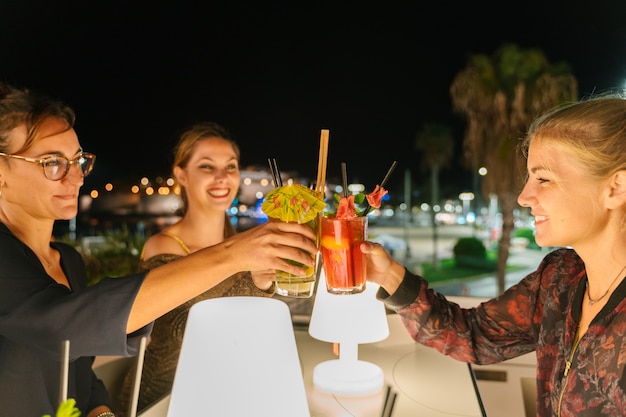 Selektiver Fokus auf drei junge Frauen, die nachts auf einer Terrasse einen Toast mit Cocktails machen