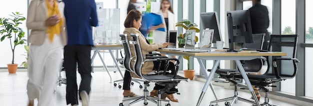 Selektiver Fokus auf eine Geschäftsfrau, die auf Laptops am Konferenztisch sitzt und arbeitet, während andere verschwommene Geschäftsfrauen in Eile im Büro mit mehreren Glasfenstern spazieren.