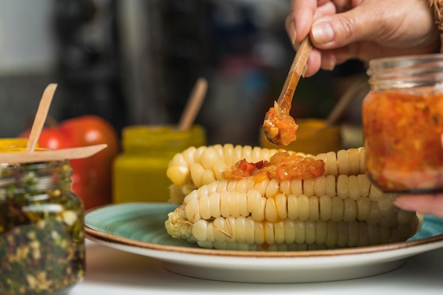 Selektiver Fokus auf eine Hand, die Rocoto-Sauce mit einem Holzlöffel auf einen gedämpften Mais in einer Küche verteilt