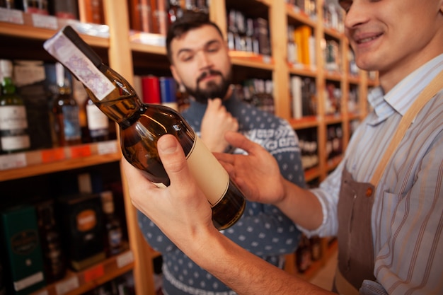 Selektiver Fokus auf eine Weinflasche in den Händen eines Winzers im Gespräch mit einem Kunden. Hübscher Mann, der leckeren alternden Wein am Supermarkt kauft