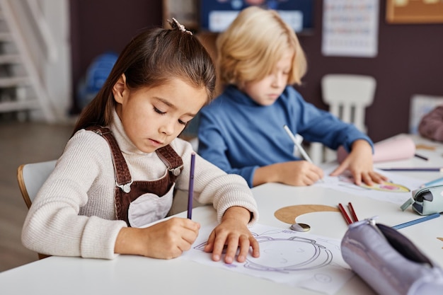 Selektiver Fokus auf fleißige hübsche Grundschüler, die im Sitzen mit Buntstiften auf Papier zeichnen