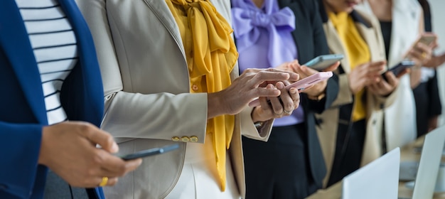 Selektiver Fokus auf fünf asiatische Geschäftsfrauen in formellen Anzügen, die im Büro mit Smartphones zusammenstehen.