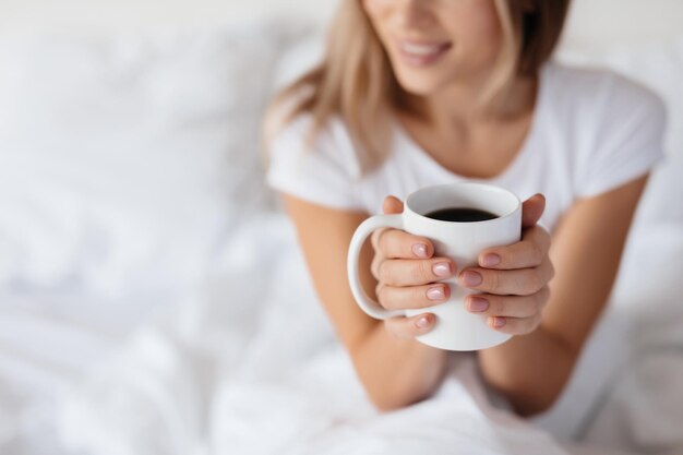 Selektiver Fokus einer weißen Kaffeetasse, die von einer entspannten jungen Dame gehalten wird, die in ihrem Bett chillt